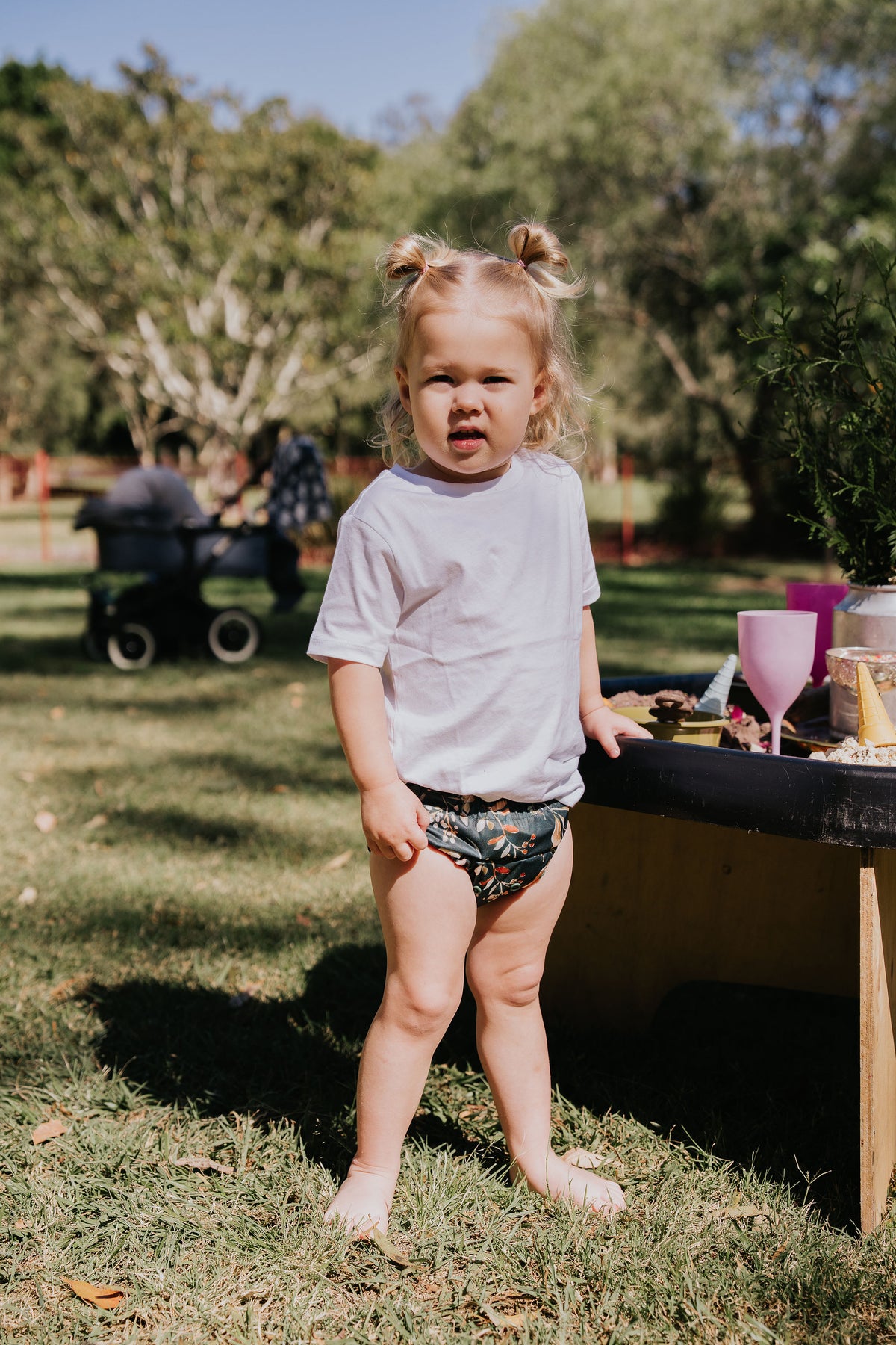 Toddler standing by a play table outdoors wearing a Gremlin pull up nappy in Evergreen print