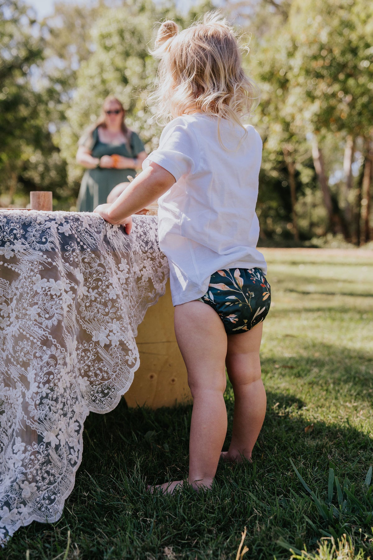 Toddler standing at a lace-covered table outdoors wearing a Gremlin pull up nappy in Evergreen print