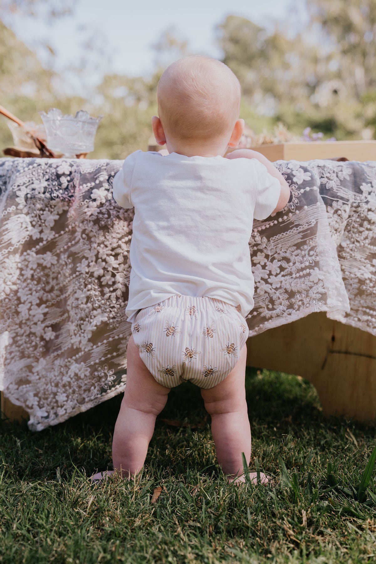Baby standing at a lace-covered table outdoors wearing a Pixie cloth nappy in Beeline print