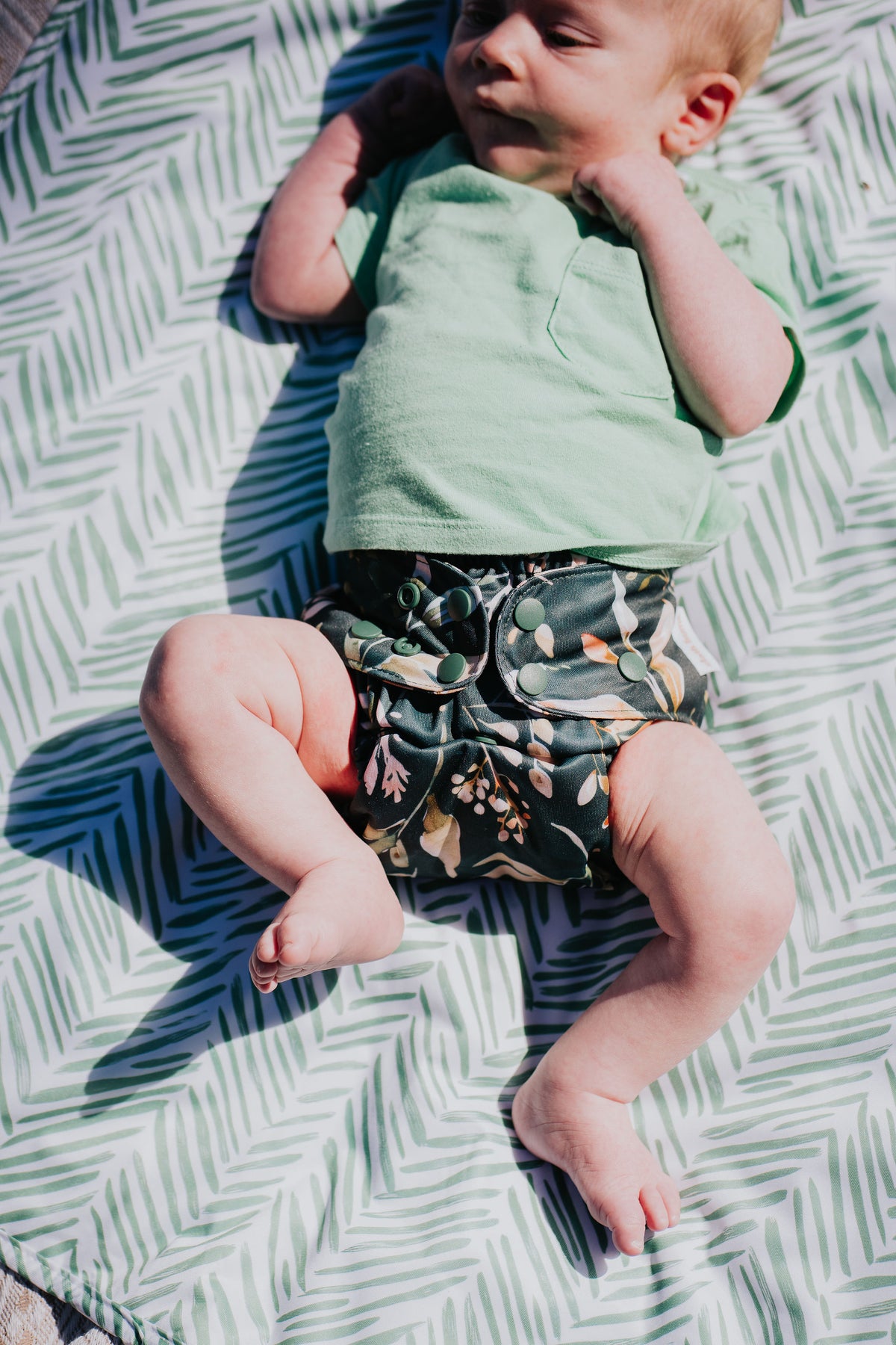 Baby lying on a leaf-print mat wearing a Pixie cloth nappy in Evergreen print