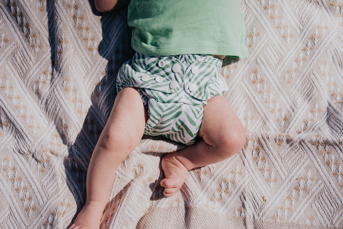 Baby lying on a picnic blanket wearing a Pixie cloth nappy in Greenhouse print