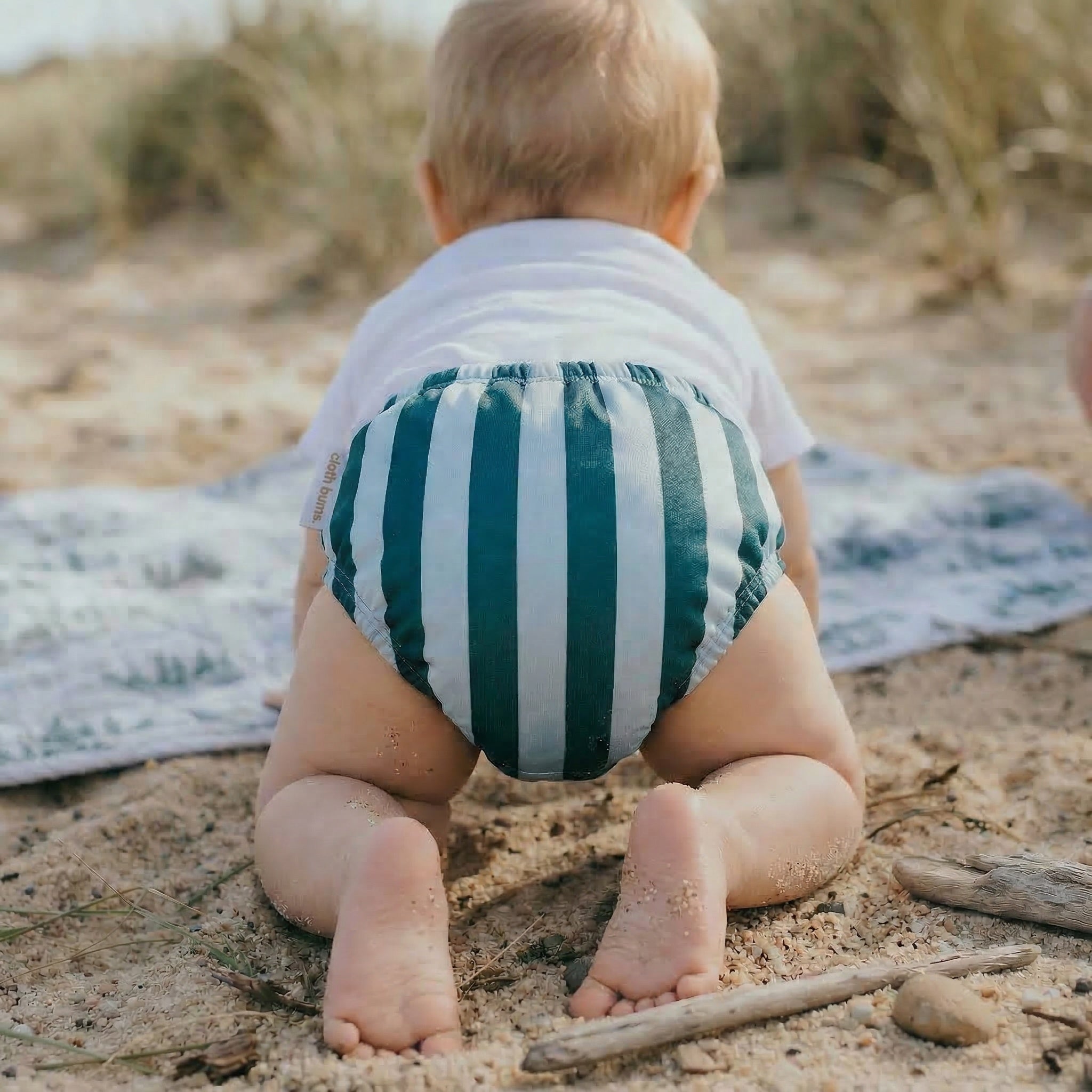 Baby sitting on the sand wearing a green and white striped diaper.